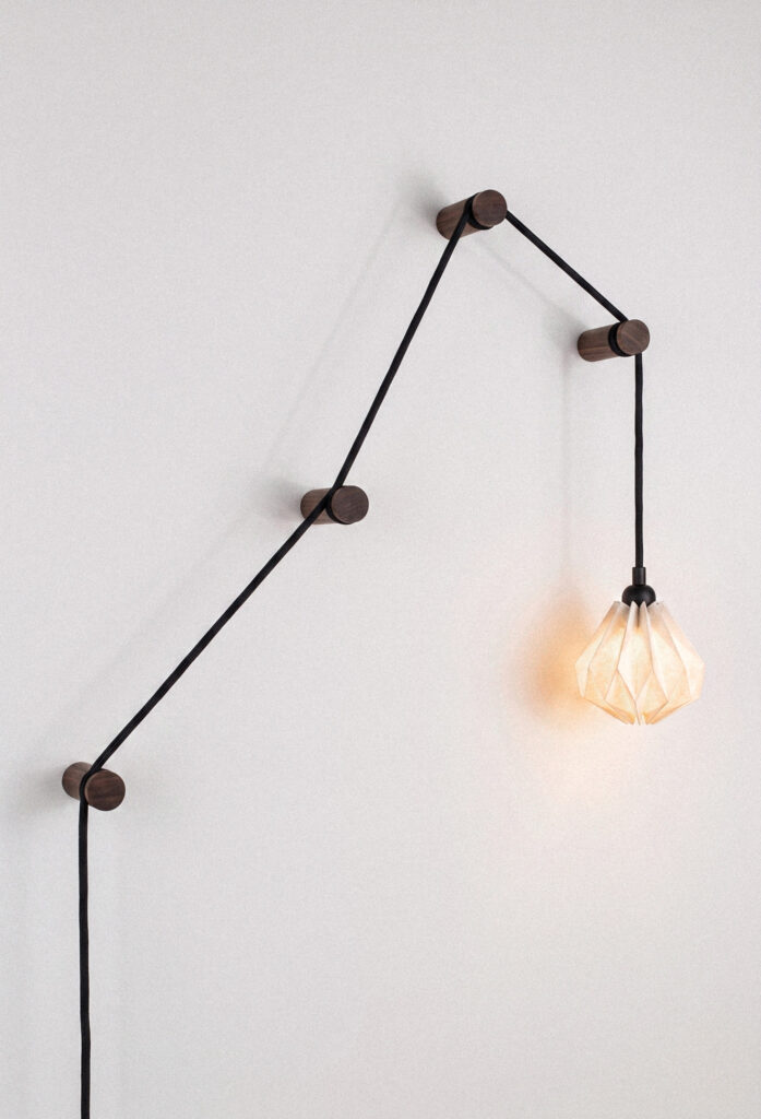 Nowoczesna, minimalistyczna lampa zamontowana na białej ścianie. Czarny przewód zasilający jest poprowadzony zygzakiem przez cztery okrągłe, ciemnodrewniane uchwyty ścienne. Na końcu kabla zwisa mały, geometryczny klosz z jasnego papieru w stylu origami, który emituje delikatne, ciepłe światło.